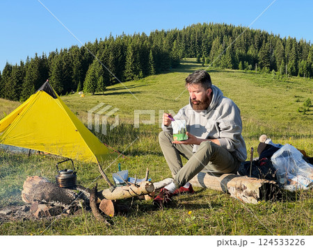 Man tourist eating from food pouch with green spoon, sits on log near campfire. Yellow tent on green hillside, surrounded by lush trees. Peacefulness of camping in beautiful natural environment. 124533226