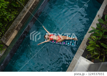 Enjoying sunbathing. Tropical vacation concept. Top view of young woman on inflatable mattress in swimming pool with palm trees. Enjoying sunbathing. Tropical vacation concept. Top view of young woman on inflatable mattress in swimming pool with palm trees. 124537031