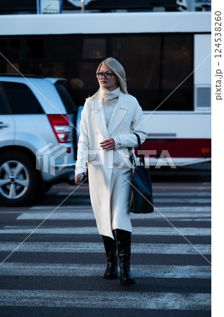 Walking confidently in the city, a fashionable woman in a long white coat crosses the crosswalk as vehicles await her stylish presence during a vibrant late afternoon Walking confidently in the city, a fashionable woman in a long white coat crosses the crosswalk as vehicles await her stylish presence during a vibrant late afternoon 124538260