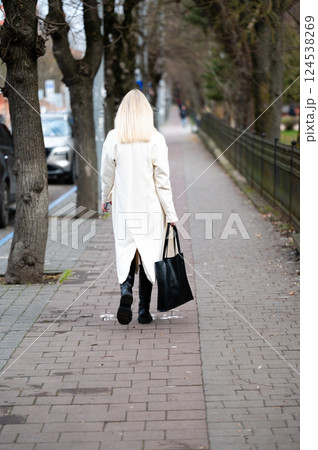 A stylish individual walks confidently along a tree-lined sidewalk, carrying a sleek black bag on a crisp autumn day while leaves rustle gently in the breeze A stylish individual walks confidently along a tree-lined sidewalk, carrying a sleek black bag on a crisp autumn day while leaves rustle gently in the breeze 124538269