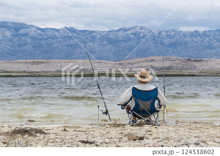 Rear view of old unrecognizable fisherman wearing a straw hat, sitting in fishing chair with two fishing poles. 124538602
