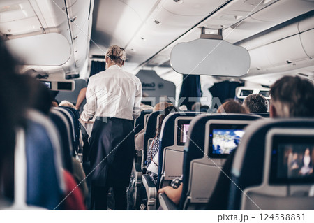 Interior of airplane with passengers on seats and stewardess in uniform walking the aisle, serving people. Commercial economy flight service concept 124538831