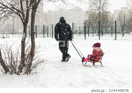 A father pulls sled with a child on a frosty cold winter day. A walk with a little kid in wintry season. Falling snowflakes in urban scene. Family fun 124540676