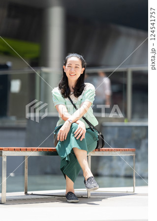 Aa Asian stylish woman with long hair and green blue dress is sitting there pleasantly grinning. Aa Asian stylish woman with long hair and green blue dress is sitting there pleasantly grinning. 124541757