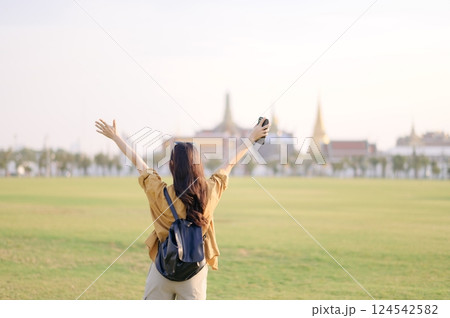 A Traveler Asian woman in 30s, bathed in golden glow of a Bangkok sunset, laughs with carefree joy, her arms outstretched as if to embrace freedom of the moment. Backpack strapped on. A Traveler Asian woman in 30s, bathed in golden glow of a Bangkok sunset, laughs with carefree joy, her arms outstretched as if to embrace freedom of the moment. Backpack strapped on. 124542582