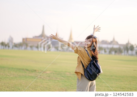 A Traveler Asian woman in 30s, bathed in golden glow of a Bangkok sunset, laughs with carefree joy, her arms outstretched as if to embrace freedom of the moment. Backpack strapped on. 124542583