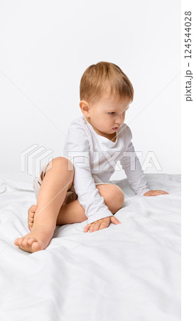 Focused little boy sitting on bed with curious look against white studio background. 124544028