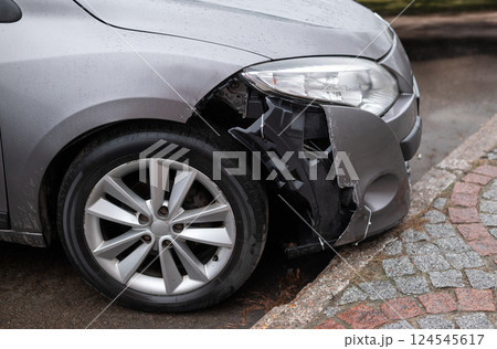Front of a silver car with a damaged bumper and exposed inner structure after an accident, parked near a curb on a wet road Front of a silver car with a damaged bumper and exposed inner structure after an accident, parked near a curb on a wet road 124545617
