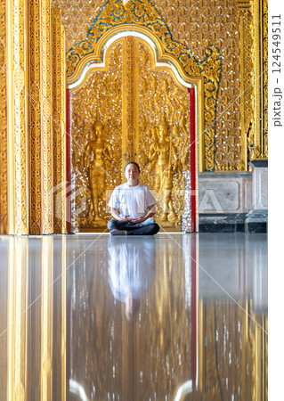Asian Man meditates on the ground with reflection in the sunlight pouring into the temple church with gold gold Thai pattern blur background. 124549511