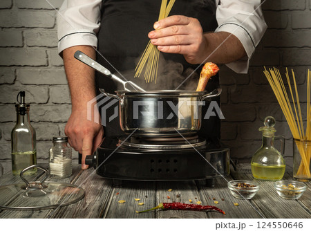 A chef prepares to cook pasta by adding dry spaghetti to boiling water in a metal pot. Surrounding ingredients include olive oil, spices, and herbs on a rustic table A chef prepares to cook pasta by adding dry spaghetti to boiling water in a metal pot. Surrounding ingredients include olive oil, spices, and herbs on a rustic table 124550646