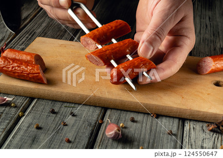 A person uses a skewer to hold sliced sausages while cooking. The setting includes wooden surfaces and scattered spices, indicating meal preparation in a cozy environment 124550647