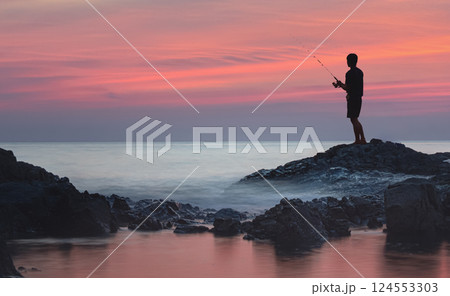 silhouette of a male fisherman above the foam waves. He is spinning fishing from the coastal stone ridge in Khao Lak. The water is filled with red glare from the shining sunset 124553303
