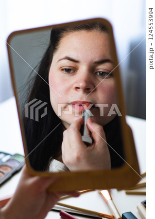 Vertical image of Millennial Caucasian woman doing self make up applying lipstick to lips while looking in mirror. Reflection in mirror. Getting ready. Beauty concept. 124553994