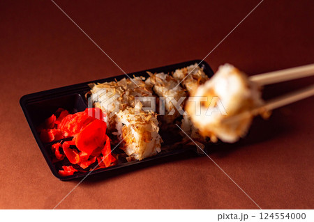 Chopsticks taking portion of sushi roll. Sushi rolls with salmon cream cheese and cucumber in take away package. Food delivery concept. 124554000