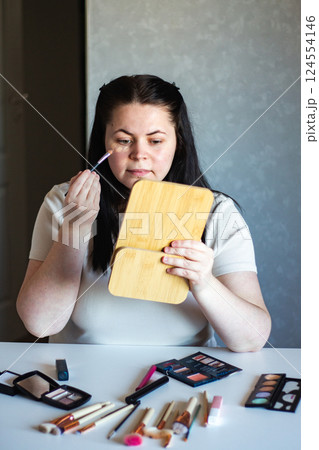 Vertical image of Millennial Caucasian woman doing self make up applying concealer on face with make up brush while looking in mirror. Getting ready. Beauty concept. 124554146