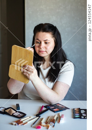 Vertical image of Millennial Caucasian woman doing self make up applying concealer on face with make up brush while looking in mirror. Getting ready. Beauty concept. Vertical image of Millennial Caucasian woman doing self make up applying concealer on face with make up brush while looking in mirror. Getting ready. Beauty concept. 124554147