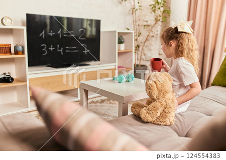 Girl watching math equations on TV with teddy bear and headphones on table, enjoying interactive online lesson. 124554383