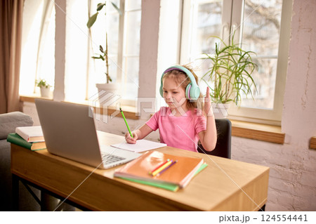 Little girl, pupil in pink shirt and headphones, sitting at desk at home, raising hand during online class while writing notes with pencil. Little girl, pupil in pink shirt and headphones, sitting at desk at home, raising hand during online class while writing notes with pencil. 124554441