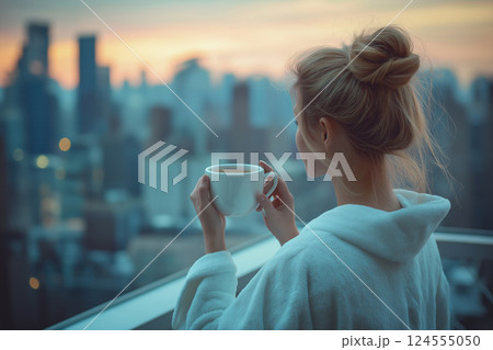 Back view of a woman standing on a city balcony, holding a morning coffee mug while wearing a fluffy white robe. Captures peaceful moments of relaxation in an urban setting during sunrise. 124555050
