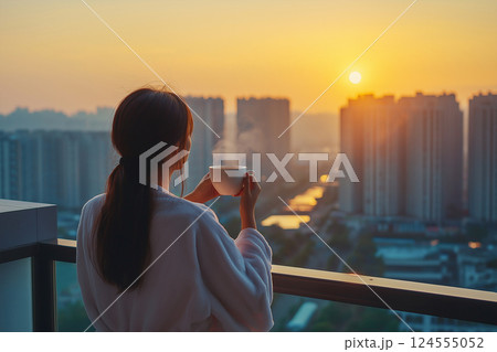 Back view of a woman standing on a city balcony, holding a morning coffee mug while wearing a fluffy white robe. Captures peaceful moments of relaxation in an urban setting during sunrise. 124555052