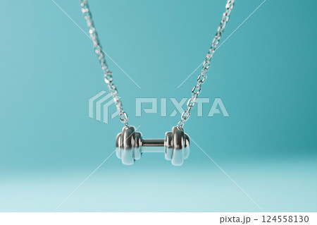 A silver chain necklace with a dumbbell-shaped pendant dangling on a blue backdrop. Close-up of a silver necklace as a symbol of fitness, exercises and sport. No excuses, stop wishing, start doing. A silver chain necklace with a dumbbell-shaped pendant dangling on a blue backdrop. Close-up of a silver necklace as a symbol of fitness, exercises and sport. No excuses, stop wishing, start doing. 124558130