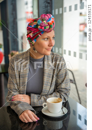 Stylish young woman in a turban sitting in a cafe, drinking coffee and enjoying the moment. The concept highlights that cancer patients can express their identity through fashion and live fully 124559171