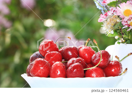 A cup of cherries on a sunny table in a flowered garden, selective focus. Sweet cherry with droplets and a branch of lilac in the background in the garden, the effect of natural bokeh in the A cup of cherries on a sunny table in a flowered garden, selective focus. Sweet cherry with droplets and a branch of lilac in the background in the garden, the effect of natural bokeh in the 124560461