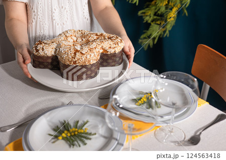 Close-up of a woman's hands serving the traditional Italian Easter cake, Colomba, on a festive table with elegant tableware and mimosa, creating a spring holiday atmosphere. 124563418