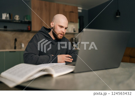 Focused bald young man in hoodie taking notes sitting at kitchen table with laptop computer, managing online business and enjoying comfort of working from home. Concept of remote job, technology. 124563432