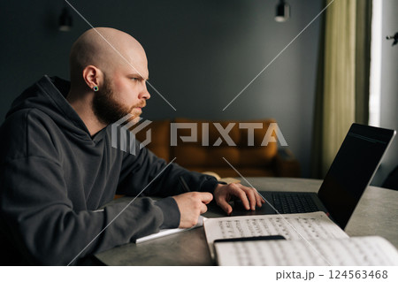 Side view of talented composer writing music notes on paper sitting at table with laptop, books, sheet music in home office, immersing in creativity and inspiration. Concept of remote job, technology. Side view of talented composer writing music notes on paper sitting at table with laptop, books, sheet music in home office, immersing in creativity and inspiration. Concept of remote job, technology. 124563468