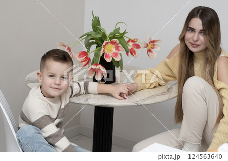 Concept family. Beautiful 19 year old girl and 5 year old boy sitting charmingly at the round table with a bouquet of red tulips, on a white background. Brother and sister bond. 124563640
