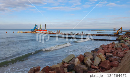 Construction machinery carries out shore protection works on stormy Baltic sea coast by laying large boulders and driving piles at sunny winter day. With no people seascape. Construction machinery carries out shore protection works on stormy Baltic sea coast by laying large boulders and driving piles at sunny winter day. With no people seascape. 124564471