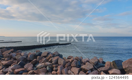 Empty rocky coast of Baltic sea with calm waves rolling on wooden breakwater and large boulders as shore protection under cloudy sky at overcast winter day. With no people desolate seascape. 124564495