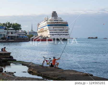 fisherman is fishing on the pier with a cruise ship in the background. International seaport. Batumi. Caught fish in the sea with a line fisherman is fishing on the pier with a cruise ship in the background. International seaport. Batumi. Caught fish in the sea with a line 124565201