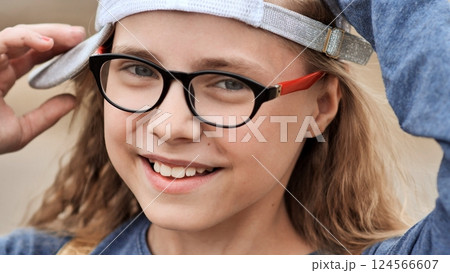 Cheerful school girl wearing glasses, smiling brightly while adjusting her cap, radiating joy and confidence in a natural outdoor setting Cheerful school girl wearing glasses, smiling brightly while adjusting her cap, radiating joy and confidence in a natural outdoor setting 124566607