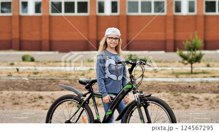 Schoolgirl leaning on bicycle near school entrance, smiling during bright daytime moment 124567295