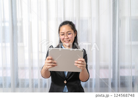 Smiling Asian Woman holds and watches gray tablet on her hand in front of curtain in private room in Work from Home period. Smiling Asian Woman holds and watches gray tablet on her hand in front of curtain in private room in Work from Home period. 124568748
