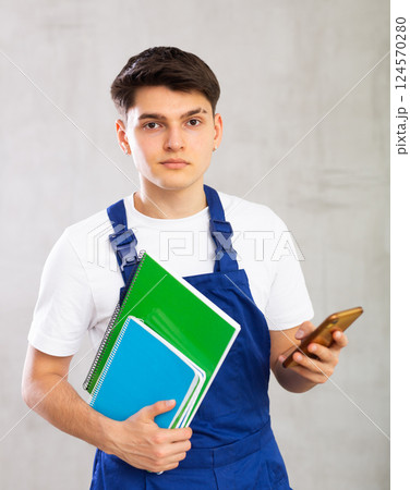 Young male builder in protective overalls with notebooks in his hands writes a message on smartphone screen 124570280