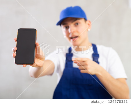 Unfocused man in jumpsuit and cap shows close up empty phone screen and points at device with finger 124570331