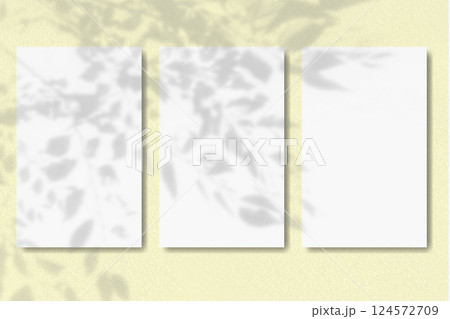 Natural light casts shadow from the leaves of a tree branch on 3 vertical sheets of textured white paper on yellow table background. Mockup Natural light casts shadow from the leaves of a tree branch on 3 vertical sheets of textured white paper on yellow table background. Mockup 124572709