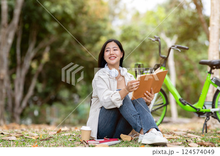 A woman looking happily at her notebook as she figure something out while sitting on park sidewalk 124574049