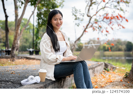 A woman looking happily at her notebook as she figure something out while sitting on park sidewalk 124574100