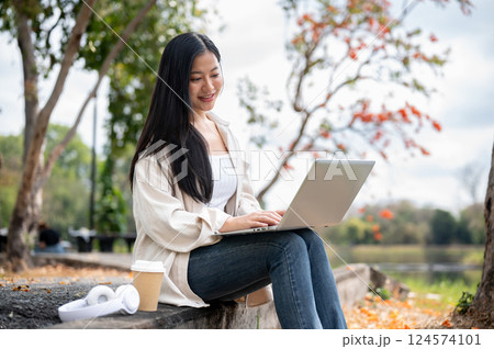 A woman looking happily at her notebook as she figure something out while sitting on park sidewalk 124574101