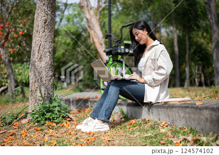 A woman looking happily at her notebook as she figure something out while sitting on park sidewalk 124574102