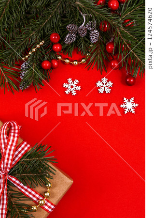 Christmas decorative border with green spruce, holly berries, snowflakes, cones and giftbox on red background, top view, vertical image, copy space. 124574620