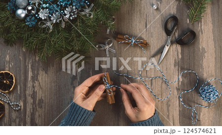 Making Christmas decoration, process. Female hands with cinnamon sticks next to blue Christmas wreath, scissors, twine on a wooden table, flat lay. Making Christmas decoration, process. Female hands with cinnamon sticks next to blue Christmas wreath, scissors, twine on a wooden table, flat lay. 124574621
