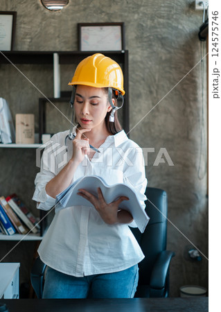 Female engineer wearing a hard hat examines blueprints in a contemporary office, highlighting construction and planning. 124575746