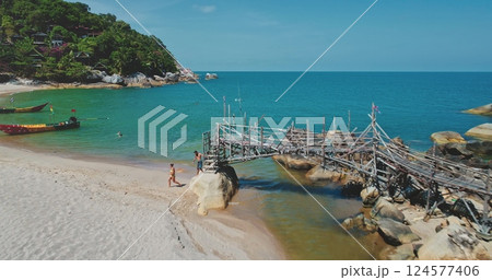 Aerial drone footage capturing a picturesque tropical beach with tourists strolling along the shore, a rickety wooden pier over turquoise waters, and lush greenery on the hillside 124577406