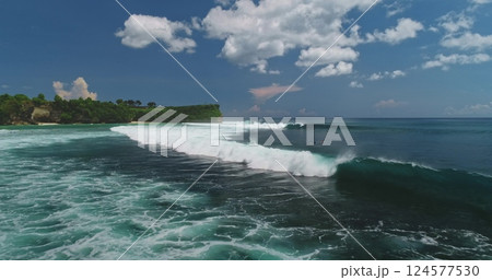 Stunning aerial view capturing turquoise ocean waves breaking on Dreamland Beach in Bali, framed by lush green cliffs and white clouds, creating a tropical paradise 124577530