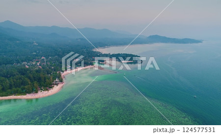 Aerial view of a tropical island coastline showing crystal clear turquoise water, lush green vegetation, boats sailing, and mountains in the distance 124577531